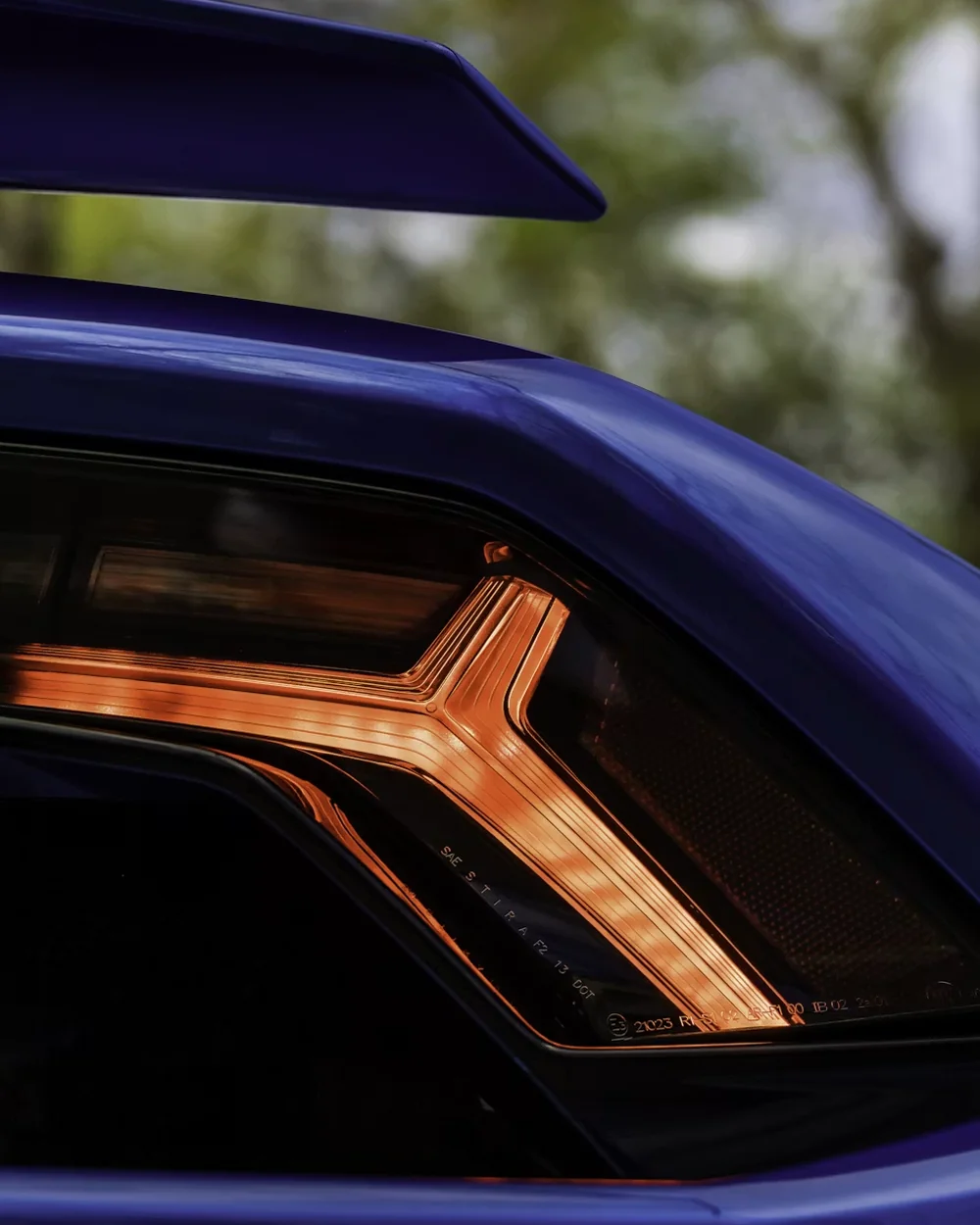Close-up of a blue car's illuminated taillight and rear spoiler in daylight