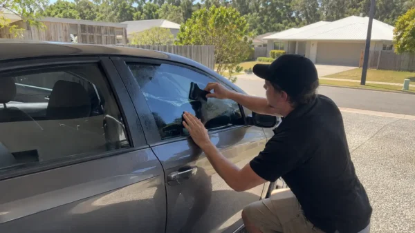 Mobile window tinting technician applying tint film to a car side window in a suburban Brisbane driveway.